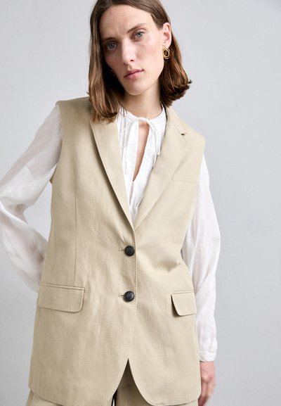 Gilet beige ajusté avec une coupe adaptée, deux poches avant et boutons noirs. Associé à une chemise blanche à manches longues et à boutons.