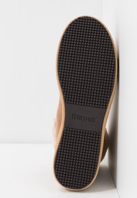 Semelle de chaussure couleur fauve avec grip en caoutchouc noir texturé et logo "Blauer USA", positionnée à la verticale contre des murs et un sol blancs.