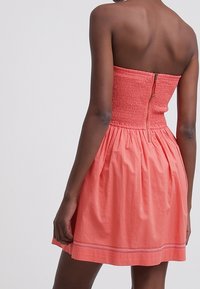 Korálově červené šaty bez ramínek s nařaseným živůtkem a rozšířenou sukní. Mají zip na zadní straně a jemný detail na lemu.