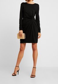 Svart långärmad klänning med knutdetalj framtill. Matchad med en texturerad beige väska och svarta sandaletter med remmar. Slät tygfinish.