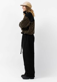 Luipaardprint fleecejack met een hoge kraag, gecombineerd met zwarte relaxed-fit broek. Glimlachend figuur met een beige pet en zwarte schoenen.