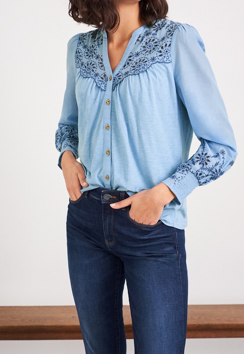 Femme portant un chemisier bleu clair avec broderie florale sur le yoke et les manches, associé à un jean bleu foncé, mains dans les poches.