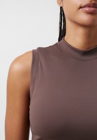 Brun ärmlös topp med hög krage, slät textur och synliga sömnadsdetaljer på axlarna, med en figurnära silhuett.