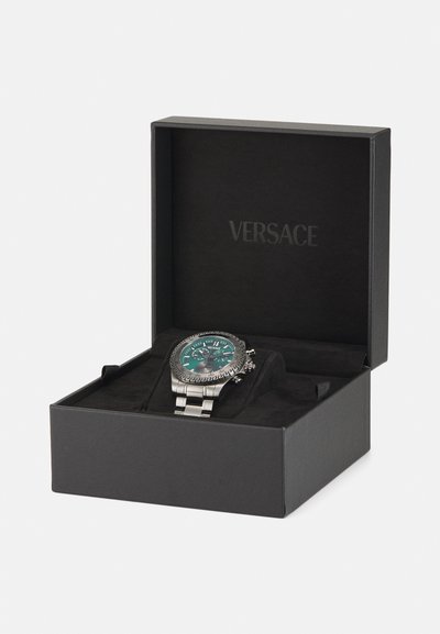 Versace watch in a black box features a teal dial, silver stainless steel band, textured bezel, and prominently displayed hour markers.