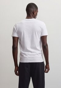 Uomo in piedi di spalle alla fotocamera, indossa una semplice t-shirt bianca e pantaloni neri, su uno sfondo chiaro e uniforme.