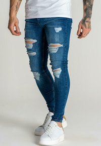 Jeans skinny desgastados en azul con rasgaduras, hechos de mezclilla. Combinados con zapatillas blancas con acentos texturizados y suelas de color claro.