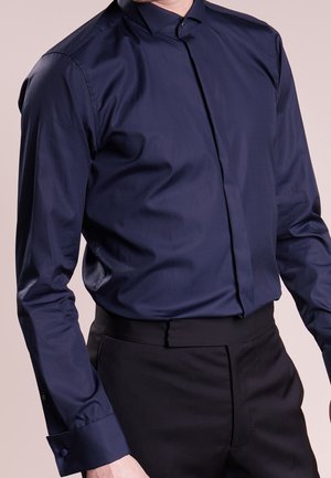 Mężczyzna w ciemnogranatowej koszuli włożonej do czarnych, dopasowanych spodni, stojący na tle jednolitego jasnego tła.