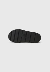 Black rubber shoe sole with a textured surface, featuring horizontal ridges and a central logo detail. The shape is rounded at the toe.