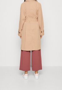 Beżowy trencz z kołnierzem i paskiem w talii, z zapinanymi na guziki rękawami, w połączeniu z szerokimi bordowymi spodniami i białymi trampkami.