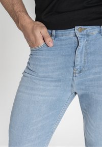 Jeans de mezclilla azul claro con un corte ajustado, que cuentan con bolsillos delanteros, costuras doradas y un cierre de botón metálico en la cintura.