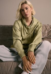 Nadrozmerná mikina s kapucňou v olivovo zelenej farbe so šnúrkami, rebrovanými manžetami, doplnená o svetlosivé tepláky, sediaca na mäkkej pohovke.