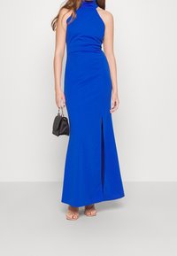 Niebieska, bezrękawnikowa sukienka maxi z wysoką szyją, dopasowanym gorsetem i bocznym rozcięciem. Modelka trzyma czarną torebkę i nosi cieliste sandały. Gładka tekstura materiału.