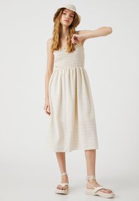 Vaaleanbeige midi-pituinen mekko, jossa on rypytetty yläosa, liehuva hame ja hienovarainen rakenne. Yhdistetty valkoisiin tasapohjaisiin sandaaleihin ja yhteensopivaan ämpärilakkiin.