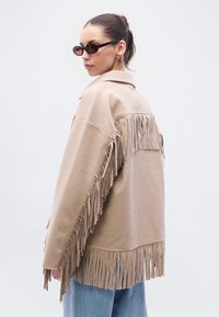 Beežika tikkudega jakk krae, pikkade varrukate ja tekstuurse kangaga, siniste teksade ja tumedate päikeseprillidega.