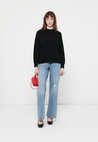 Femme se tenant contre un fond blanc, vêtue d'un pull noir, d'un jean déchiré bleu clair, portant des escarpins noirs, tenant un petit sac à main rouge dans la main gauche.