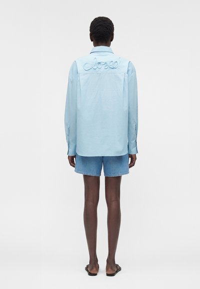 Άτομο που φοράει ανοιχτό μπλε μακρυμάνικο πουκάμισο με κεντημένη τη λέξη "closed" στην πλάτη, τζιν σορτς και μαύρα σανδάλια, που στέκεται με την πλάτη σε εμάς.