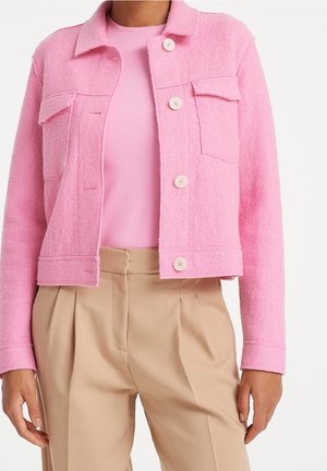 Mujer que lleva una chaqueta de lana color rosa claro con botones sobre una camisa rosa y pantalones plisados beige de talle alto, frente a un fondo blanco.
