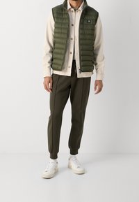 Man som bär en olivgrön puffväst, enfärgad beige skjorta, mörkgröna joggingsbyxor, vita strumpor och vita sneakers, stående på vit golv.