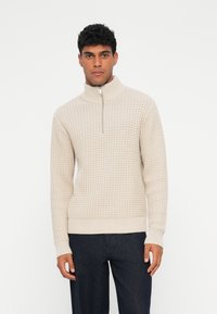 Beige quarter-zip genser med teksturet mønster, ribbestrikkede mansjetter og kant. Kombinert med mørke bukser som har høy krage-design.