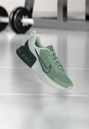 Baskets de sport vertes et blanches avec le swoosh noir de Nike et une semelle à coussin d’air visible, placées sur un sol de gym réfléchissant avec des poids en arrière-plan.