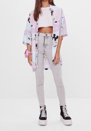 Vrouw draagt een oversized pastel T-shirt met Mickey Mouse-print, witte cropped top, lichtgrijze high-waisted jeans, zwarte platformsneakers en een roze polszakje.