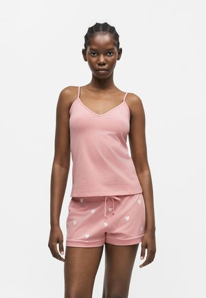 Ung kvinde iført pink ærmeløs top og matchende pink shorts med hvide hjerte mønstre, stående mod en ensfarvet hvid baggrund.