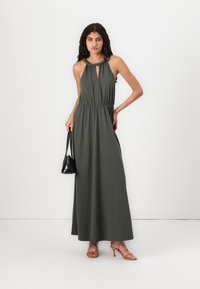 Vestido maxi em verde escuro, sem mangas, com detalhe de recorte, cintura elástica e material fluido. Combinado com sapatos de salto finos e uma pequena bolsa preta.