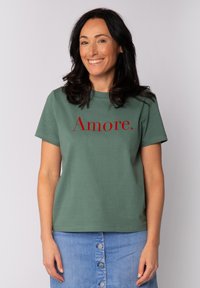 T-shirt en coton vert avec le texte imprimé rouge "Amore" sur le devant, col rond et manches courtes. Associé à une jupe en denim.