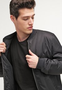 Chaqueta bomber negra con cierre frontal, puños y cuello acanalados, con una textura suave y brillante. Llevada sobre una camiseta negra.