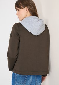 Femme portant une veste marron foncé avec une capuche gris clair, tournée de côté montrant le dos de la veste et les détails de la capuche.