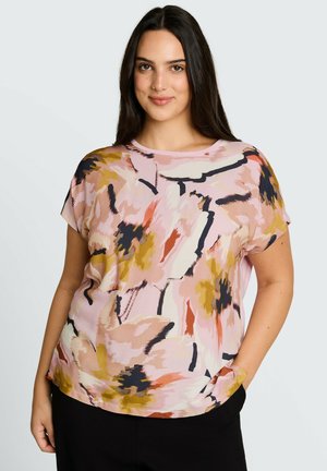 Femme souriante aux longs cheveux foncés portant un chemisier rose à manches courtes avec des motifs floraux abstraits jaunes, noirs et orange et un pantalon noir.