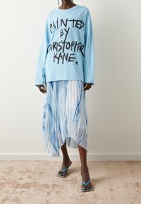 Himmelblaues Langarmhemd mit schwarzem Text, kombiniert mit einem plissierten, transparenten Rock mit blauen Batikmustern und blauen Flip-Flops.