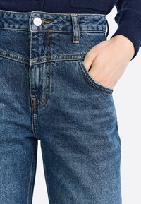 Jeans de mezclilla en azul oscuro con cintura alta, que cuentan con cinco bolsillos, costuras visibles en naranja y un botón de metal en la cintura.
