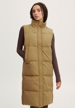 Gilet rembourré kaki avec col haut, fermeture éclair à l'avant et poches latérales. Présente une texture matelassée et une silhouette longue jusqu'aux genoux.