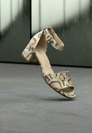 Ανοιχτόχρωμο μπεζ και καφέ σανδάλι με μοτίβο φολιδωτού δέρματος, ανοιχτές μύτες, λουράκι στον αστράγαλο και χαμηλό χοντρό τακούνι πάνω σε γκρι πάτωμα με θολό φόντο.