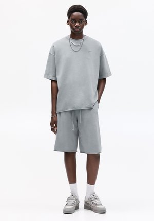 Lys grå oversized kortærmet sweatshirt med matchende shorts, der har en tekstureret overflade og snoretræk, parret med grå sneakers.
