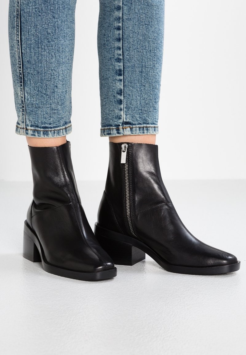 Bottes cheville en cuir noir avec zips latéraux et talons blocs portées avec un jean bleu roulé sur un sol blanc.