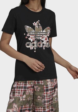 Henkilö, jolla on päällään musta Adidas-t-paita, jossa on kukkakuvioinen logo, ja siihen sopivat ruutukuosiset shortsit, joissa on kukkakuvio, kädet rennosti sivuilla valkoista taustaa vasten.