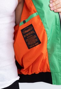 Veste bombers avec un intérieur orange et un extérieur vert. Présente des poignets élastiques, un ourlet noir et une étiquette noire avec du texte doré.