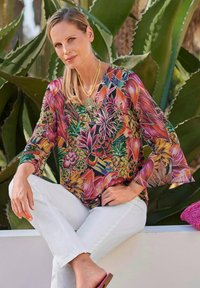 Bunte, kurzärmelige Bluse mit einem Blumen- und Früchte-Muster in Pink, Grün und Gelb. Kombiniert mit weißen Hosen in lockerer Passform.