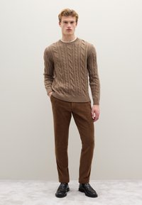 Beige Strickpullover mit Zopfmuster und langen Ärmeln, kombiniert mit braunen Cordhosen. Modell trägt schwarze Loafers, vor einem neutralen Hintergrund.