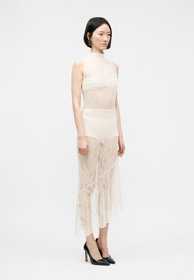 Femme aux cheveux courts et noirs porte une robe en dentelle blanche transparente sans manches par-dessus des sous-vêtements assortis et des talons hauts noirs à bouts pointus.