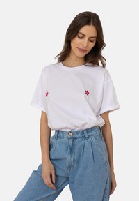 Biała bawełniana koszulka z krótkim rękawem, ozdobiona niewielkim haftem czerwonego kwiatu na piersi, połączona z niebieskimi dżinsami o wysokim stanie.