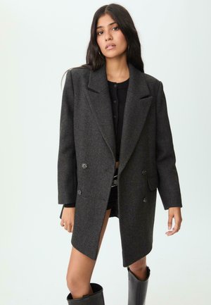 Grå dobbeltknappet blazer, lavet af tekstureret stof, har brede revers og sidelommer. Båret over en sort top og shorts.