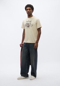 Μπεζ βαμβακερό t-shirt με γραφικό σχέδιο που περιλαμβάνει μια κορώνα και λουλουδάτα στοιχεία. Συνδυασμένο με τζιν παντελόνι φαρδύ πόδι σε μπλε χρώμα και λευκά αθλητικά παπούτσια.