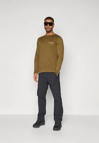 Majica od maslinasto zelene dugih rukava s logotipom, uparena s tamnim hlačama i crnim čizmama. Dodaci uključuju velike crne sunčane naočale.