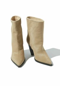 Un par de botines de ante beige con punta afilada y tacones en forma de cono negros sobre un fondo blanco que proyectan sombras.