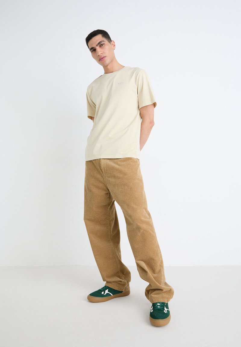 T-shirt en coton crème, coupe décontractée. Pantalons en velours côtelé marron à jambes larges. Baskets en daim vert avec des accents blancs. Fond uni.