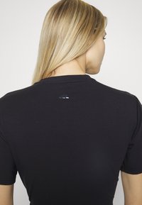 Svart kortärmad skjorta i slät tyg med en subtil lyster, med en reflekterande logotypdetalj nära halsringningen på baksidan.