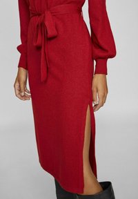 Czerwona dzianinowa sukienka zawiązywana w pasie i długimi bufkami. Posiada boczne rozcięcia i miękką teksturę. Akcentowana czarnym obuwiem.